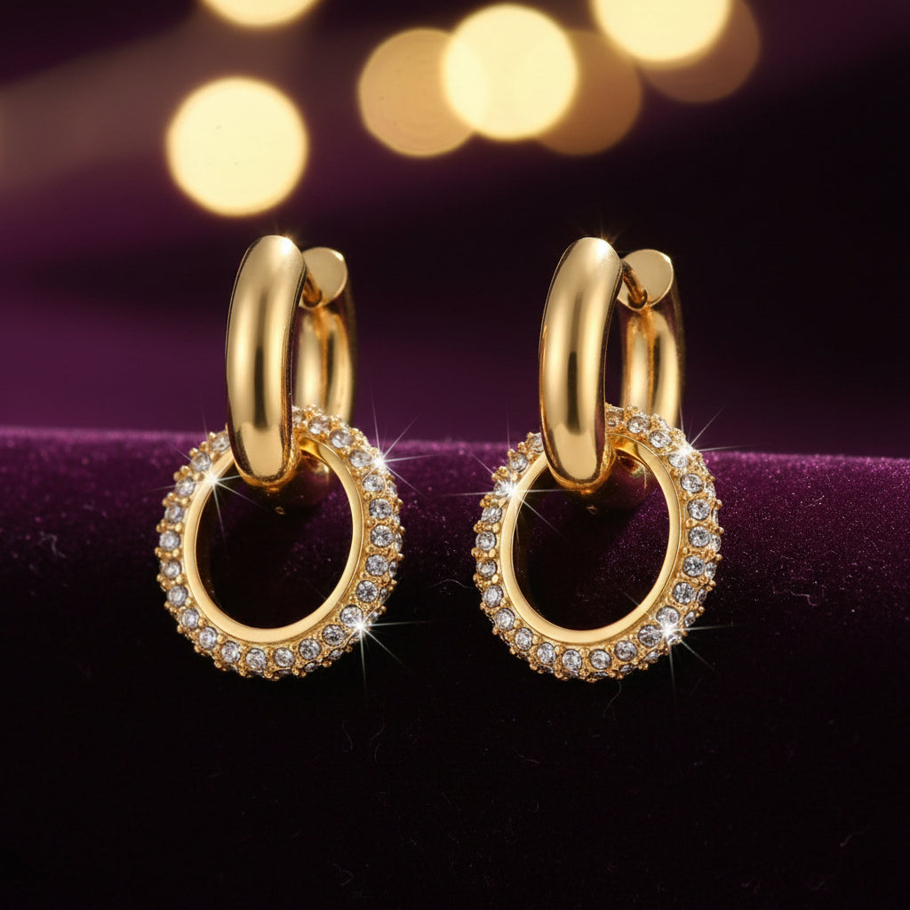 A pair of gold hoop earrings with interlocking oval drops, each adorned with a border of small clear stones, shown on a plain white background.