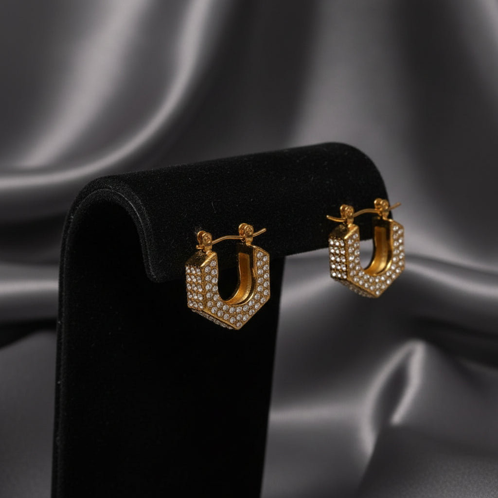 A pair of gold-toned geometric hoop earrings paved with clear rhinestones, displayed on a draped cream satin fabric background.