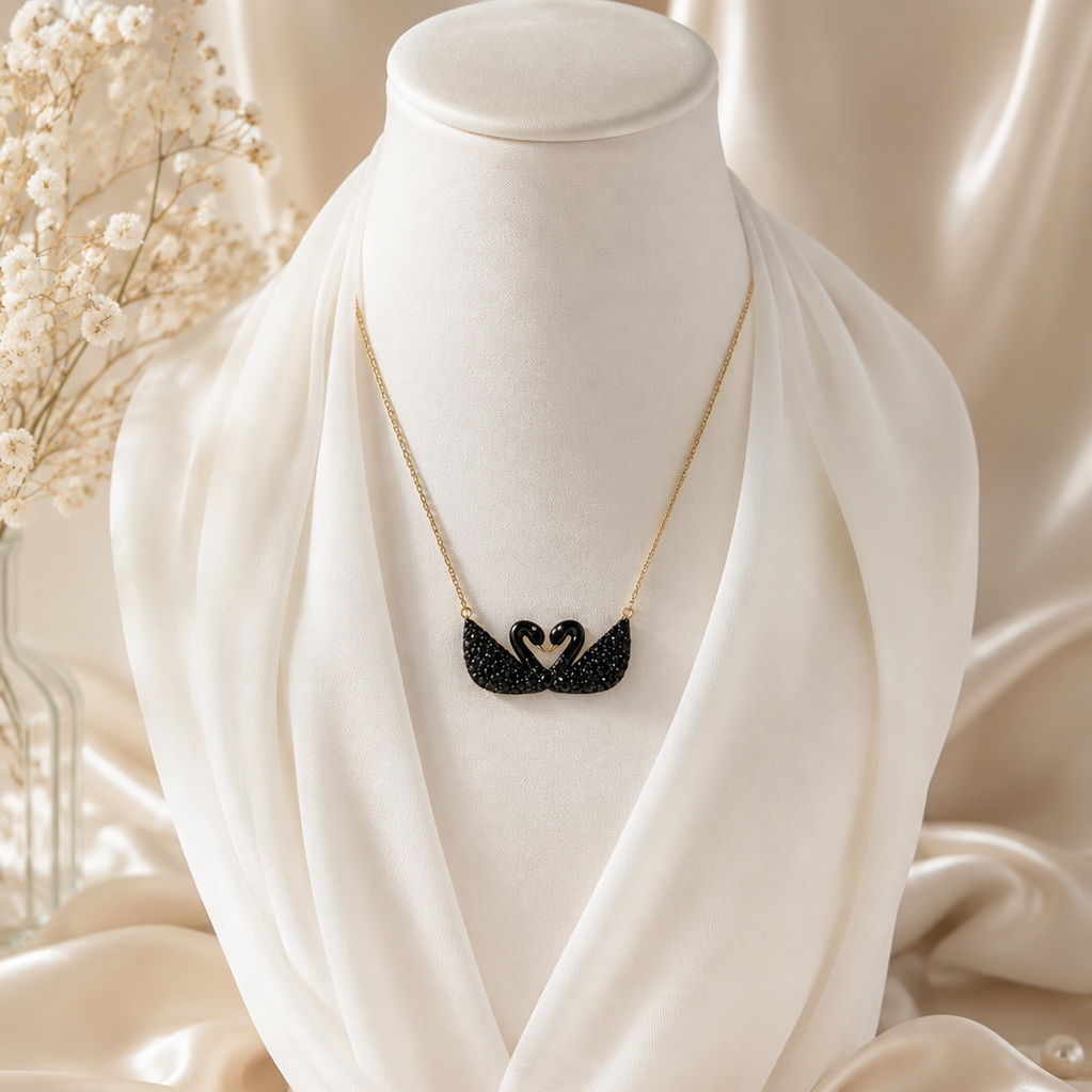A gold necklace with a black swan pendant displayed on a white fabric mannequin bust, surrounded by draped satin fabric, dried flowers, and pearl accessories. The brand name 'THE VALLEY HANI' is visible at the bottom.