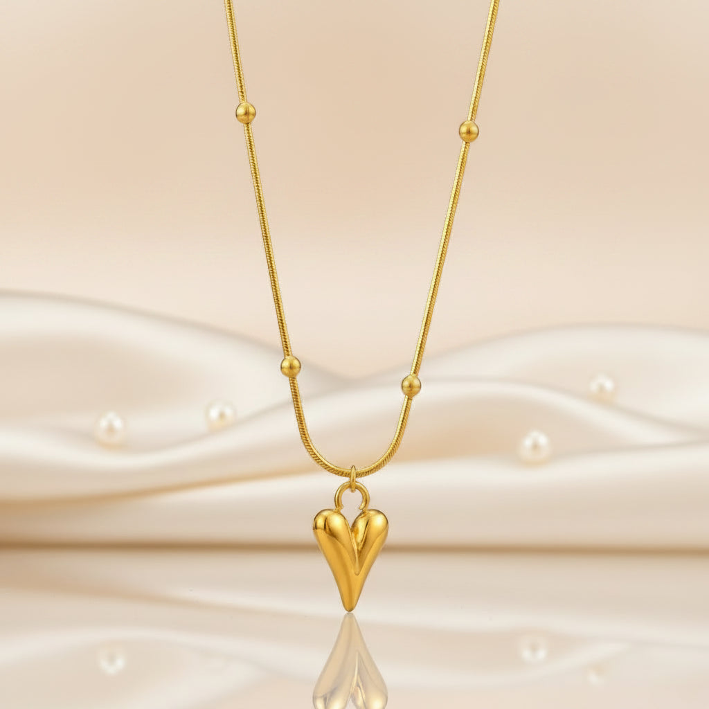 A gold necklace featuring a textured, polished heart-shaped pendant hanging from a delicate chain with small gold bead accents, set against a solid gray background.