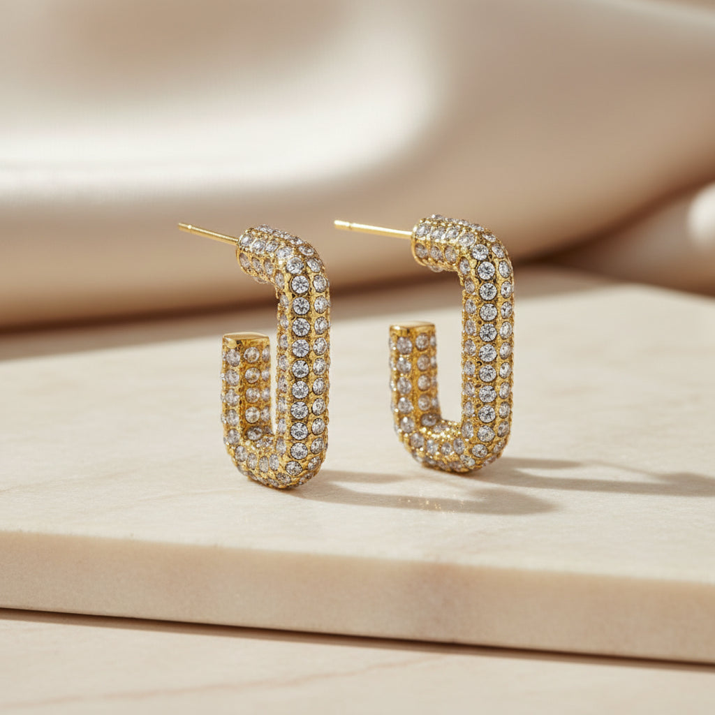 A pair of gold hoop earrings embellished with clear rhinestones, shown on a clean white background with a subtle reflection.