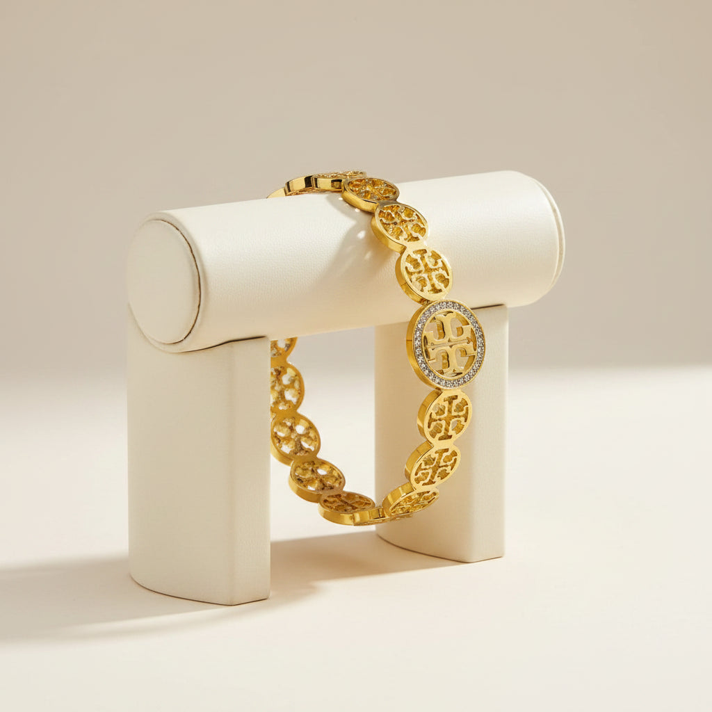 A gold-tone bracelet featuring circular interlocking monogram links, with a central oval link encrusted with small clear stones and displaying a raised monogram logo. The bracelet is photographed on a clean white background.