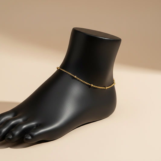 A delicate gold chain anklet with an adjustable lobster clasp, displayed on a matte black mannequin leg against a white background.